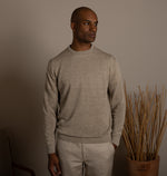 Man wearing a beige sweater and pants standing against a plain wall with a plant in the background.